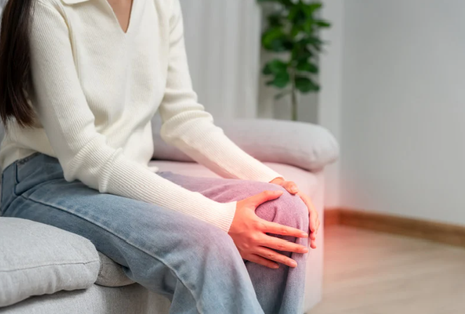 Dona asseguda al sofà agafant-se el genoll amb un efecte de dolor vermell, il·lustrant el dolor articular durant la menopausa.