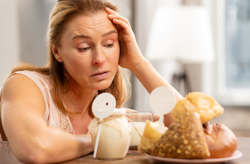 Dona madura preocupada mirant pa amb gluten i lactis en un menjador, ideal per il·lustrar la inflamació intestinal.
